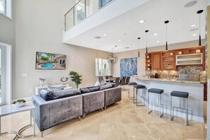 A modern kitchen with bar stools, a dining area with a gray sofa, and a large window overlooking a pool. - Luxury villa w/pool, rooftop bar & fire pit (Pompano Beach)