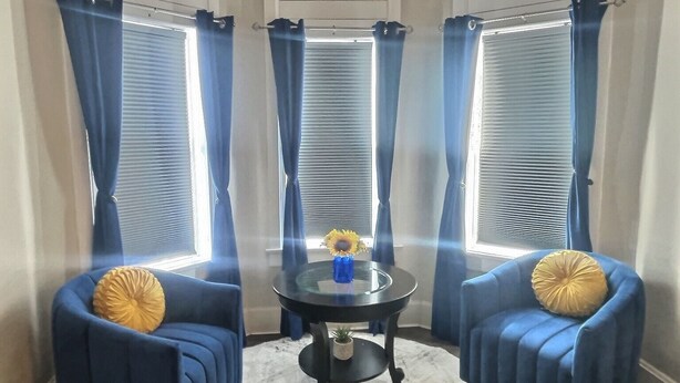Una habitación de esquina con una mesa redonda de vidrio, dos sillones azules, una alfombra blanca y una ventana con cortinas azules.