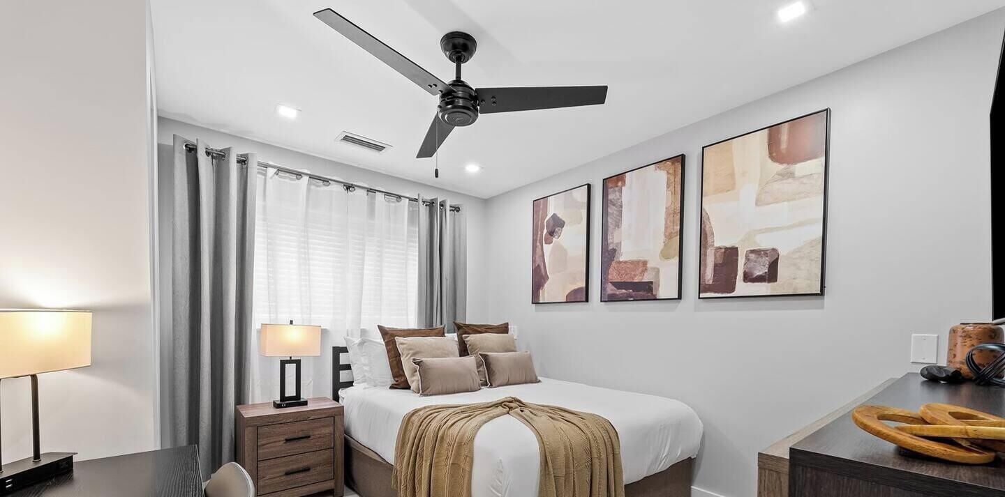 A modern bedroom with a bed, bedside table, lamp, ceiling fan, and artwork on the wall.