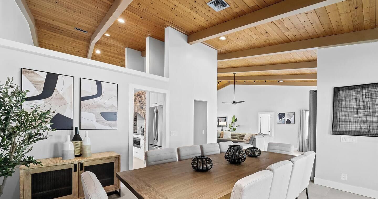 A spacious dining area with a wooden table and white chairs, a kitchen visible through an open door, and a ceiling fan.