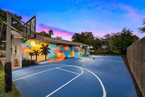 An outdoor basketball court with a colorful mural on the wall, surrounded by trees and a wooden fence. - Pool - Basketball -Soccer - Game Room - Playground (Largo)