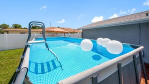 En udendørs swimmingpool med stige, omgivet af et hvidt hegn, og et hus i baggrunden.