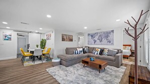 En moderne stue med en grå hjørnesofa, et træ sofabord og en grå lurvet måtte. Der er et spiseområde med et hvidt bord og gule stole.