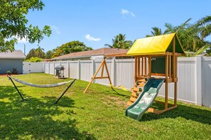 A backyard with a swing set, a hammock, and a playhouse with a slide.