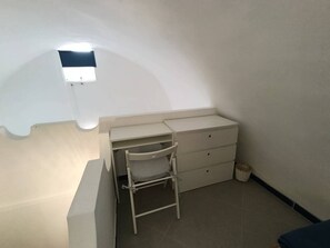Ein kleiner, weißer Raum mit Schreibtisch, Stuhl und einem kleinen Fenster.