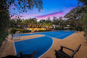 An outdoor basketball court with a net, surrounded by trees and a building with lights.