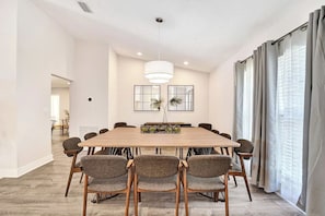 Een moderne eetkamer met een grote houten tafel, zes gestoffeerde stoelen, een hanglamp en twee spiegels aan de muur.