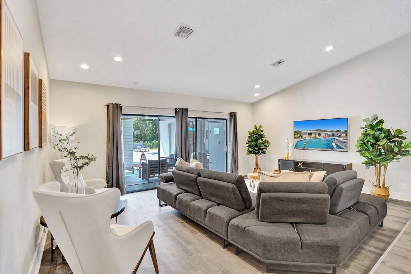 A modern living room with a grey sectional sofa, a dining table with chairs, a flat-screen TV mounted on the wall, and a large window with curtains.