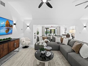 Ein modernes Wohnzimmer mit einer Eckcouch, einem Flachbildfernseher, einem runden Couchtisch mit Blumen und einem Deckenventilator.