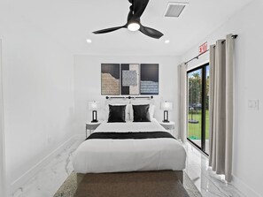 Een moderne hotelkamer met een groot bed, twee nachtkastjes met lampen, een plafondventilator en uitzicht op de omgeving door een groot raam.