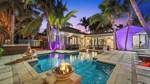 Une piscine avec un foyer, entourée de palmiers et de mobilier d’extérieur.