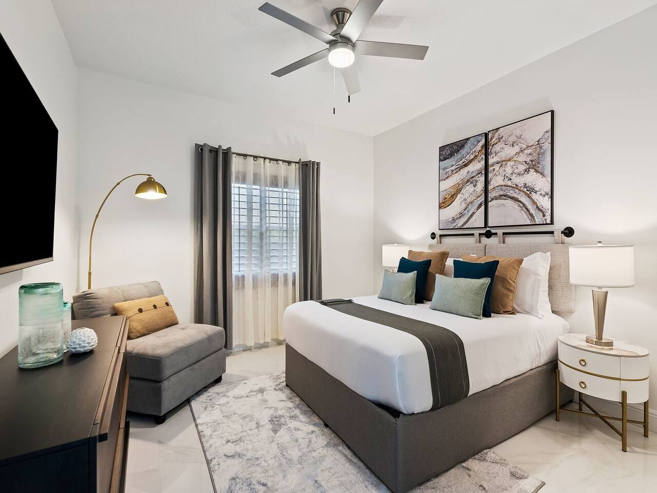 A modern hotel room with a large bed, a desk, a chair, a TV, and a ceiling fan.