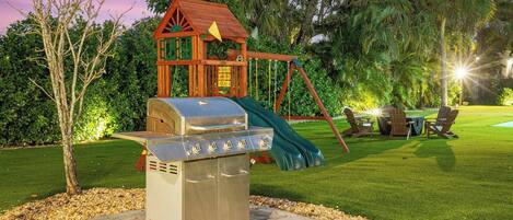 A backyard with a barbecue grill, a swing set, and a small wooden playhouse.