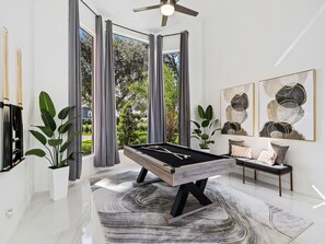 Un tavolo da biliardo in una stanza moderna con ampie finestre, una ventola a soffitto e arte astratta alle pareti.