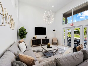 A modern living room with a large sofa, a flat-screen TV, a chandelier, and a view of the outdoors through large windows. - Villa w/pool, games & BBQ (Coral Springs)