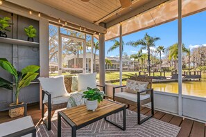 A sunlit patio with a wooden table, two white cushioned chairs, and a view of a lake and buildings in the background. - Waterfront - Pool - Pickleball - Basket -Sleeps 18 (Westchase)