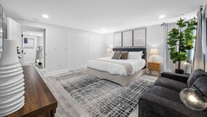 Ein modernes Schlafzimmer mit einem großen Bett, Nachttischen, einem grauen Sofa und einer Topfpflanze.