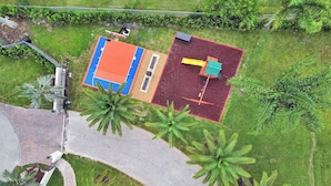 An aerial view of a property with a paved driveway, a basketball court, and a fenced area with trees.