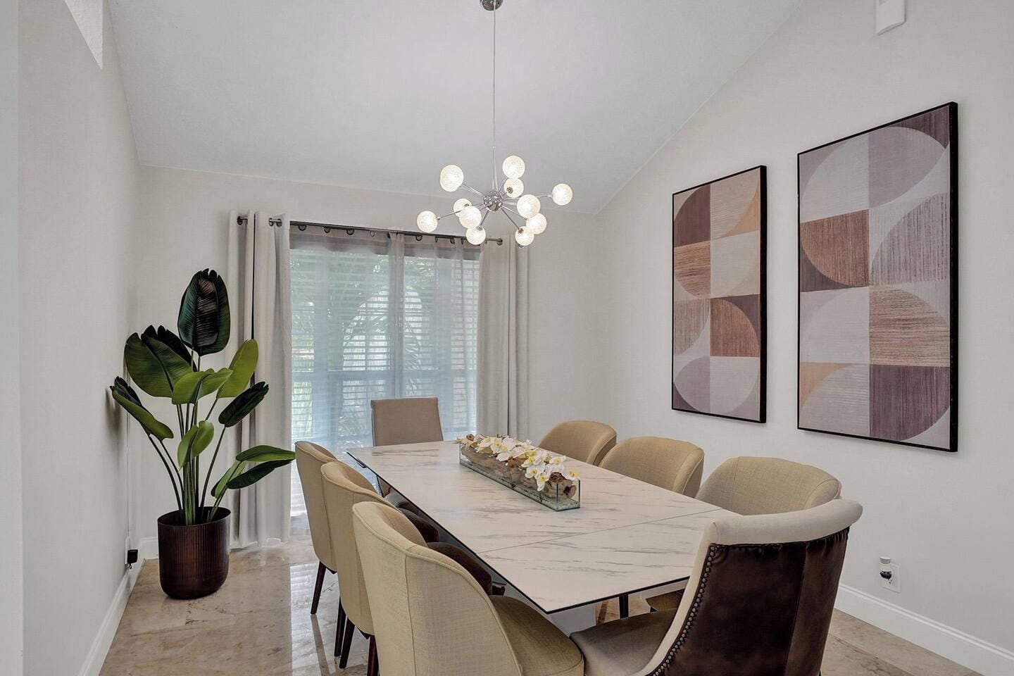 A dining area with a marble table, beige chairs, a potted plant, and abstract wall art.