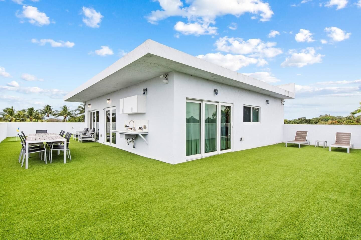 A modern white building with a flat roof, a patio with a table and chairs, and a well-maintained lawn.