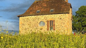 Une maison en pierre avec un toit en pente, deux fenêtres avec volets et une cheminée.