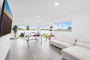 A modern living room with a white sofa, a dining table, and a large window offering a view of the outdoors. - Spacious Apartment w/Patio & Mini-Golf (Deerfield Beach)
