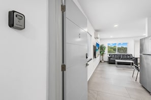 A modern interior with a white door, a black security device, a grey sofa, a table, and a refrigerator. - Spacious Apartment w/Patio & Mini-Golf (Deerfield Beach)