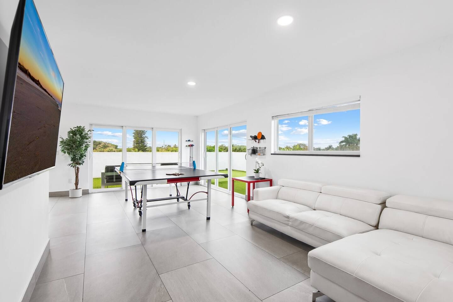 A modern living room with a white sofa, a dining table, and a large window offering a view of the outdoors.