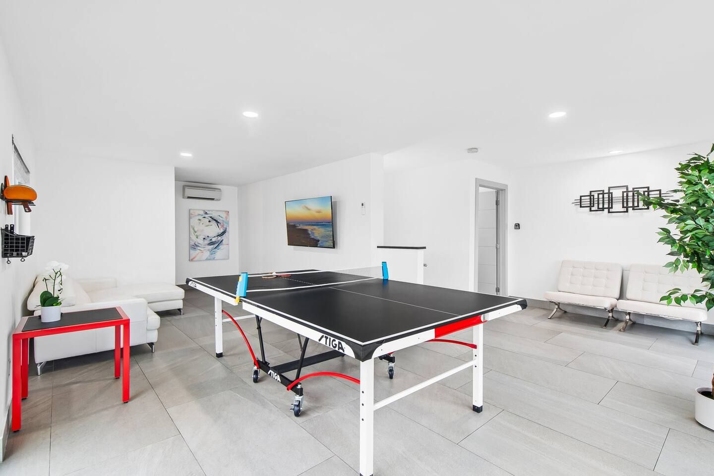 A modern room with a ping pong table, a white sofa, a red table, and a plant.