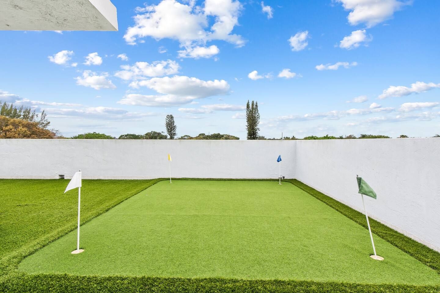Ein Golfplatz mit grüner Kunstrasen, weiß-grünen Fahnen, einer weißen Mauer und Bäumen im Hintergrund.