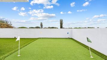 Ein Golfplatz mit grüner Kunstrasen, weiß-grünen Fahnen, einer weißen Mauer und Bäumen im Hintergrund.
