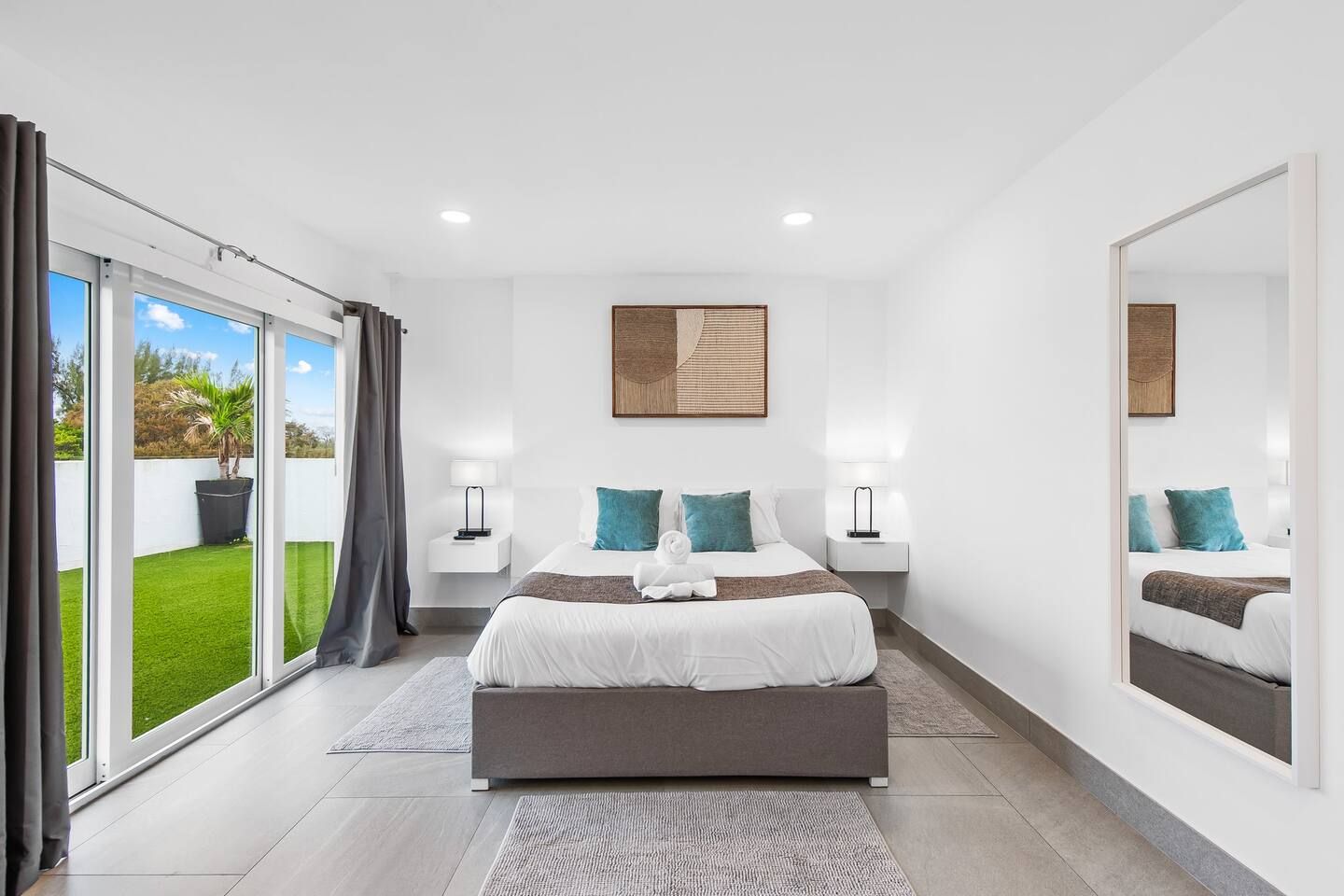 Un dormitorio moderno con una cama grande, dos mesitas de noche, un espejo y vistas a un césped verde a través de puertas correderas de vidrio.