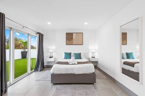 A modern bedroom with a large bed, two bedside tables, a mirror, and a view of a green lawn through sliding glass doors. - Apt w/Patio, Balcony & Mini-Golf (Deerfield Beach)