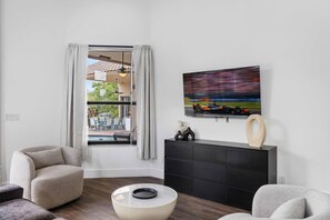 A modern living room with a TV mounted on the wall, a black entertainment unit, and a view of an outdoor patio. - Spacious home w/pool & tennis court (Sunrise)