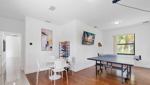 Ein Zimmer mit einem Tischtennisch, weißen Stühlen, einem Bücherregal und Kunstwerken an den Wänden.