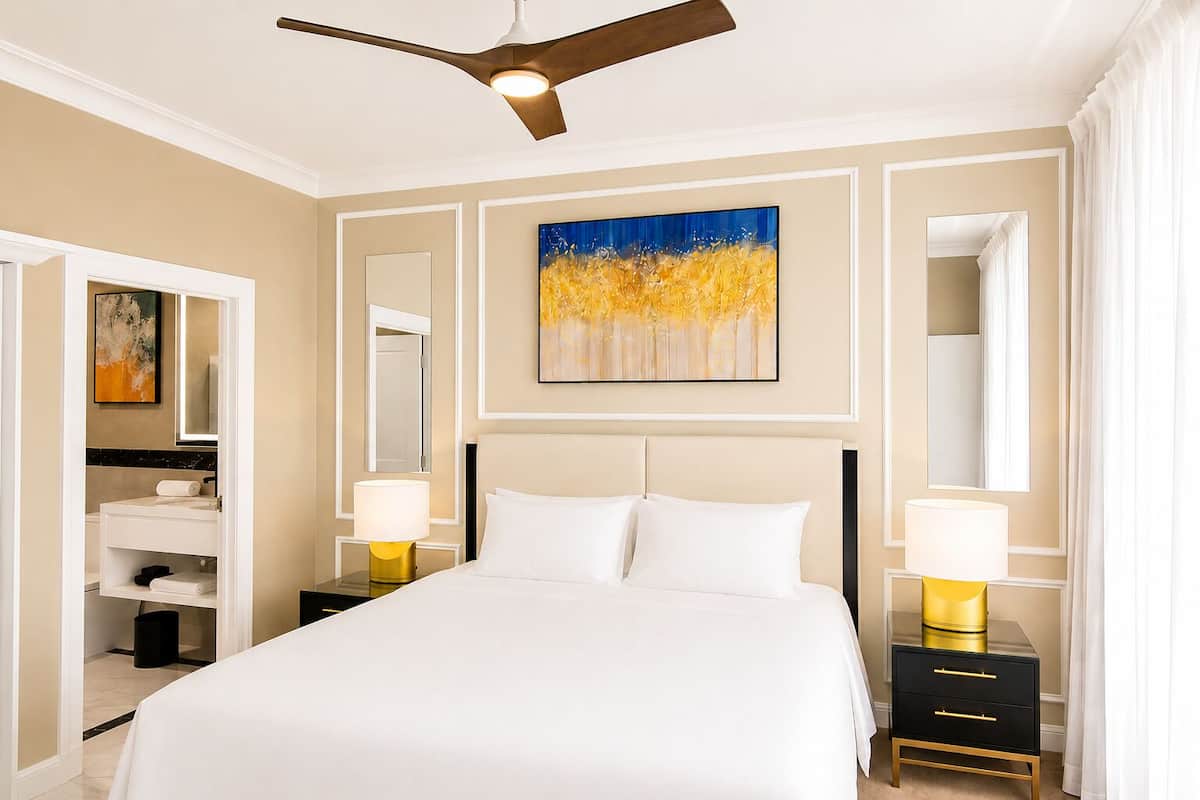 A hotel room with a bed, two bedside tables with lamps, a ceiling fan, a painting, and a bathroom with a mirror and sink.
