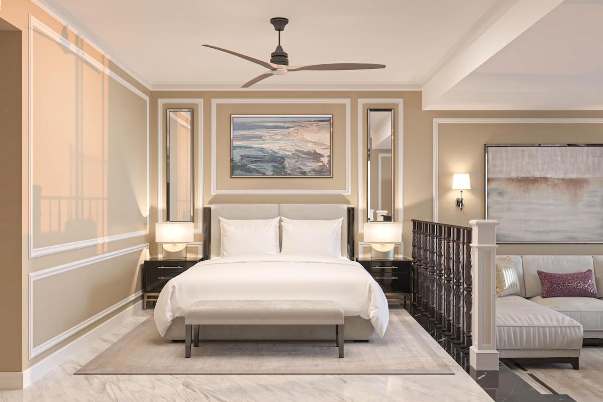 A spacious bedroom with a large bed, a ceiling fan, a painting on the wall, and a seating area with a sofa and armchair.