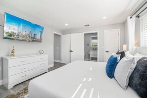 A modern bedroom with a large bed, a dresser, a mirror, and a painting of a cityscape. - Heated Pool - Putting Green - 5 min from Beach! (Deerfield Beach)