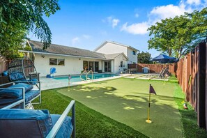 A backyard with a swimming pool, a grassy area with a putting green, and a wooden fence. - Heated Pool - Putting Green - 5 min from Beach! (Deerfield Beach)