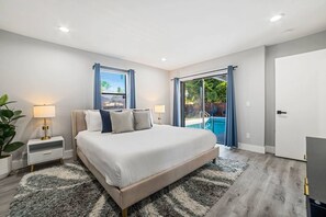 A modern bedroom with a large bed, two bedside tables with lamps, a view of a swimming pool through a sliding glass door, and a grey shaggy rug. - Heated Pool - Putting Green - 5 min from Beach! (Deerfield Beach)