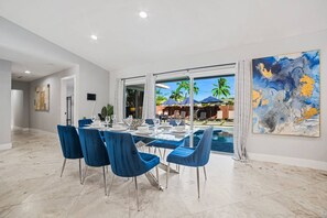 A modern dining area with a pool view, featuring a table set for six, blue chairs, and a large abstract painting on the wall. - Heated Pool - Putting Green - 5 min from Beach! (Deerfield Beach)