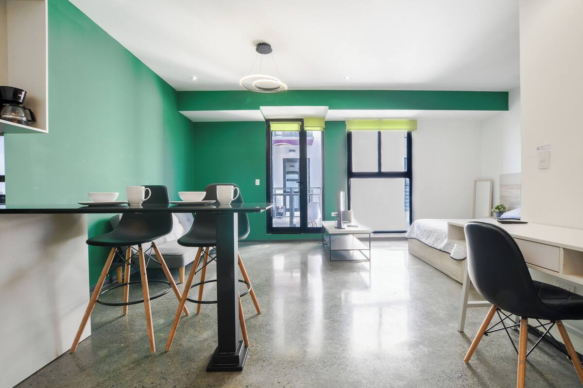 Una camera moderna con una parete verde a contrasto, un'area pranzo con tavolo e sedie, un letto e una scrivania con una sedia.