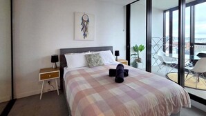 Une chambre à coucher avec un lit, des tables de chevet, une petite table, un attrape-rêves, une plante en pot et une vue sur un balcon avec des chaises.