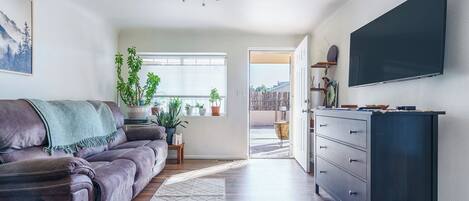 A living room with a sofa, a TV, a ceiling fan, and a view through an open door.