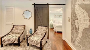 A bedroom with two patterned armchairs, a sliding barn door, a bed with white bedding, and a decorative wall. - Island Square condo #808 (Saint Simons Island)