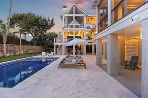 A poolside area with lounge chairs and umbrellas, a pool, and a multi-level house with outdoor lighting. - Stunning Brand NEW Build, 6 bd OCEANVIEW Home on IOP! Private pool! (Isle Of Palms)