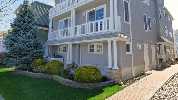 A two-story residential building with a balcony on the second floor, a well-maintained lawn, and a small evergreen tree. - Comfortable Southend 4BR Condo • Sleeps 8 • Deck, Parking & Outdoor Shower (Ocean City)