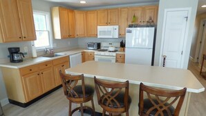 A kitchen with wooden cabinets, a white countertop, a microwave, a stove, and three wooden chairs. - Comfortable Southend 4BR Condo • Sleeps 8 • Deck, Parking & Outdoor Shower (Ocean City)