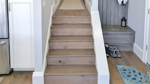 Un couloir avec un escalier en bois, un coin cuisine sur la gauche et un porte-chaussures avec des chapeaux sur la droite.
