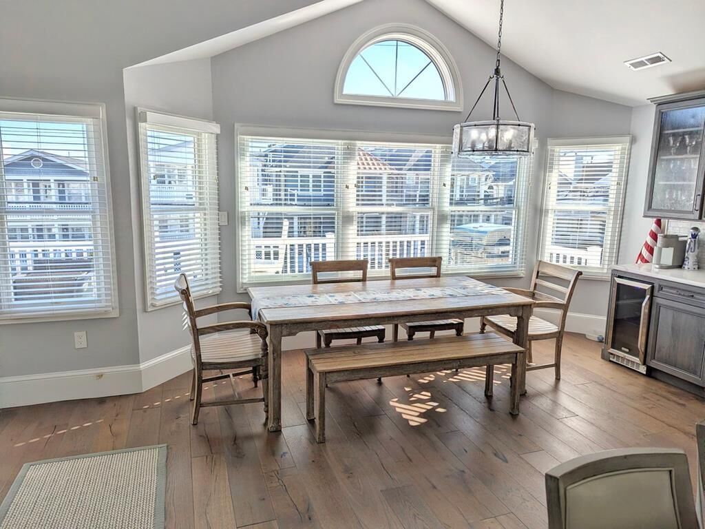 A dining area with a large wooden table and benches, surrounded by large windows with blinds, a chandelier, and a view of a building outside.